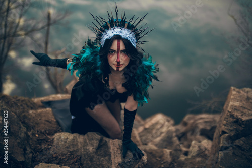 Fotografi A woman in the image of a fairy and a sorceress standing over a lake in a black dress and a crown