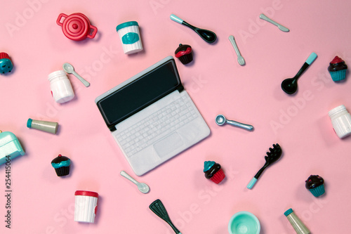 flat lay image of plastic toy tableware with cakes and laptop on pastel pink background