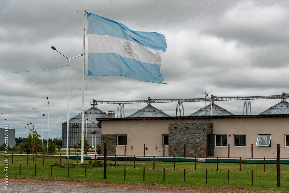 Aged Argentine flag waving in front of an industry with silos on a cloudy day