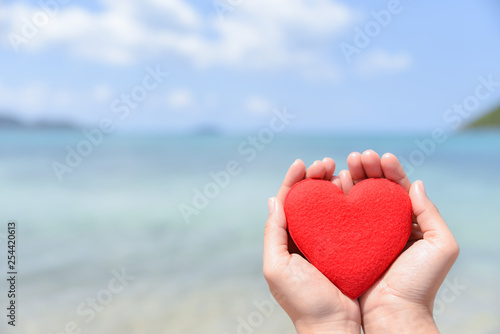 Woman hands holding a red heart on the beach with blurred sea and blue sky background. Summer day concept. Vacation and holidays concept. Love concept.