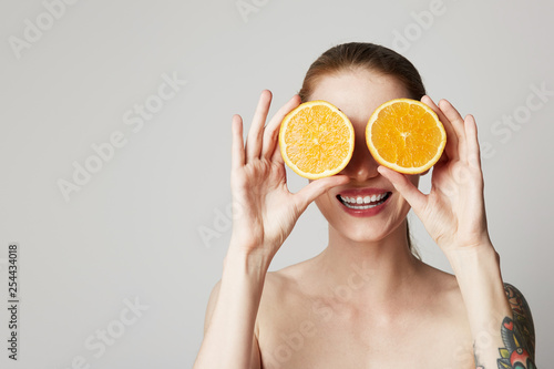 Happy cheerful tattoed young woman is holding two halves of an orange fruit in front of her eyes