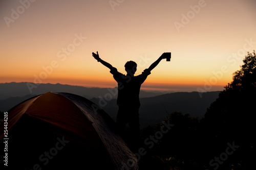 Silhouette of Happy man with holding coffee cup stay near tent around mountains under sunset light sky enjoying the leisure and freedom.