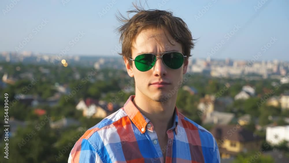 Close up portrait of young man in sunglasses sitting on rooftop of high-rise building with cityscape background. Handsome guy confident looks into camera with beautiful blurred landscape. Slow motion Close up portrait of young man in sunglasses sitting on rooftop of high-rise building with cityscape background. Handsome guy confident looks into camera with beautiful blurred landscape. Slow motion