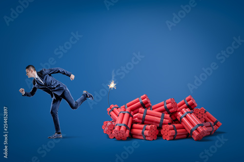 Businessman running away from pack of red tnt dynamite sticks with lighted fuse on blue background