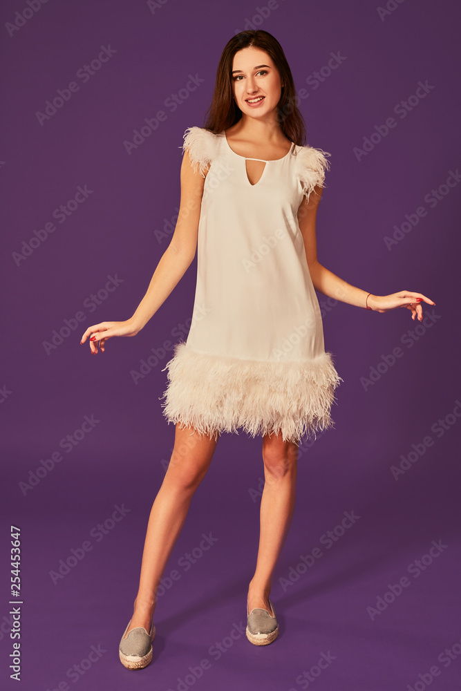 Naklejka premium Beautiful brunette woman in elegant cocktail white dress is posing in studio on purple background.