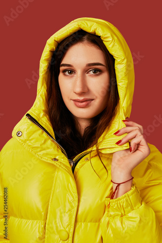 Portrait of brunette young woman wearing oversized yellow down jacket with hood on. Studio close up shot. © nazarovsergey
