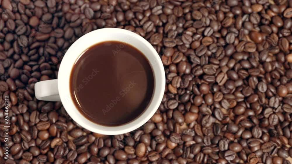 White cup (mug) filled with black hot coffe lying on brown roasted coffe beans in a café with text space on the side - top view. White cup (mug) filled with black hot coffe lying on brown roasted coffe beans in a café with text space on the side - top view.