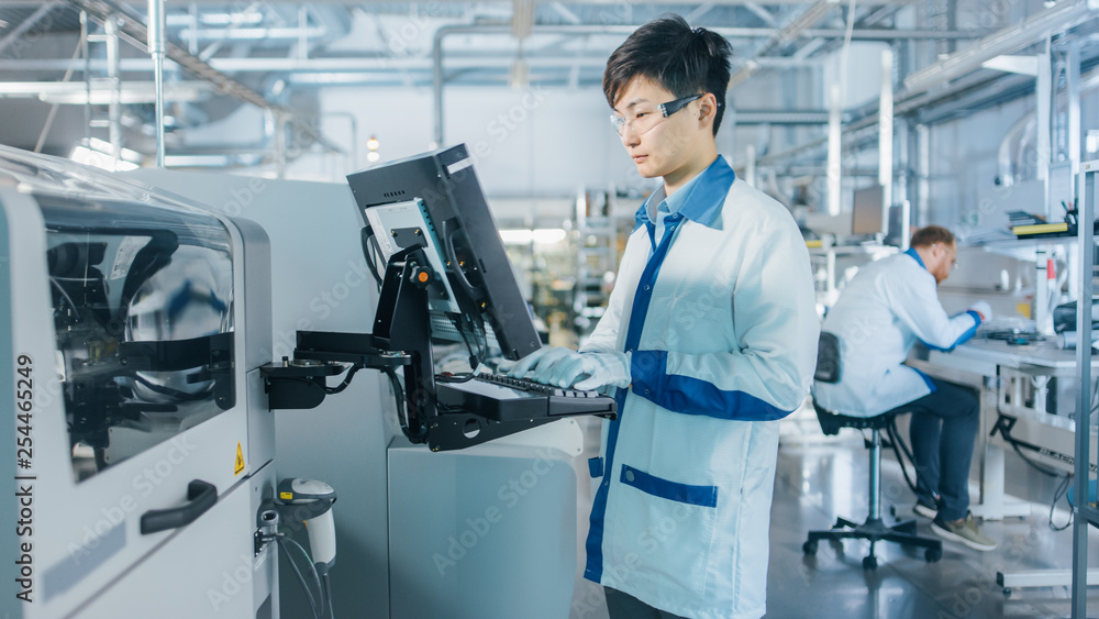 On High Tech Factory Asian Engineer Uses Computer for Programing Pick and Place Electronic Machinery for Printed Circuit Board Surface Mount Assembly Line. Production of PCB with SMT Machinery. On High Tech Factory Asian Engineer Uses Computer for Programing Pick and Place Electronic Machinery for Printed Circuit Board Surface Mount Assembly Line. Production of PCB with SMT Machinery.