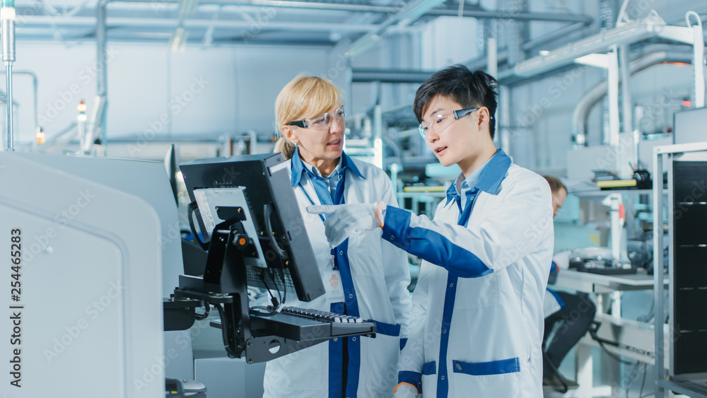 On High-Tech Factory Asian Engineer Talks with Female Supervisor, Use Computer for Programming Pick and Place Electronic Machinery for Printed Circuit Board Surface Mount Assembly Line. On High-Tech Factory Asian Engineer Talks with Female Supervisor, Use Computer for Programming Pick and Place Electronic Machinery for Printed Circuit Board Surface Mount Assembly Line.