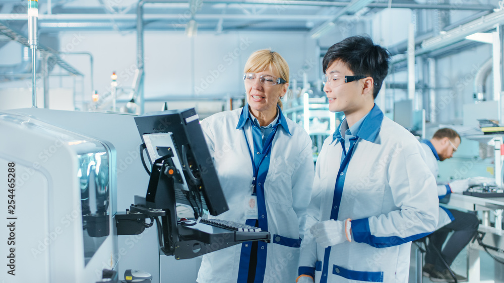 On High-Tech Factory Asian Engineer Talks with Female Supervisor, Use Computer for Programming Pick and Place Electronic Machinery for Printed Circuit Board Surface Mount Assembly Line. On High-Tech Factory Asian Engineer Talks with Female Supervisor, Use Computer for Programming Pick and Place Electronic Machinery for Printed Circuit Board Surface Mount Assembly Line.