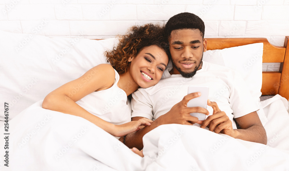 Fototapeta premium Smiling black millennial couple using smartphone on bed