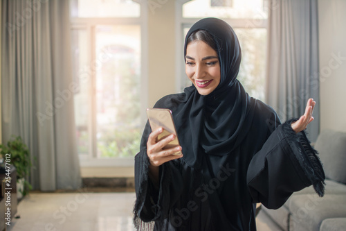 Portrait of cheerful muslim young woman in abaya using smartphone while standing in living room. photo