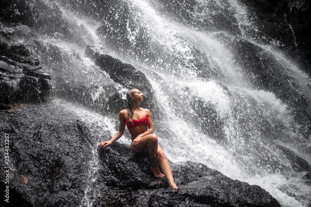 fashion portrait of beautiful woman with brunette hair in red swimwear posing  on background of waterfall in Bali