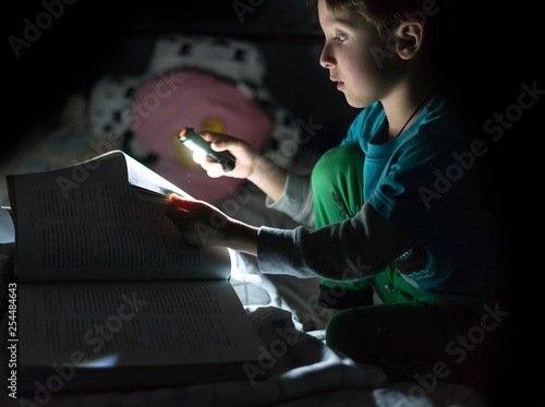 Fun caucasian boy reading a book at night with flashlight in the hand at home. Kid reading bedtime story, dark photo.