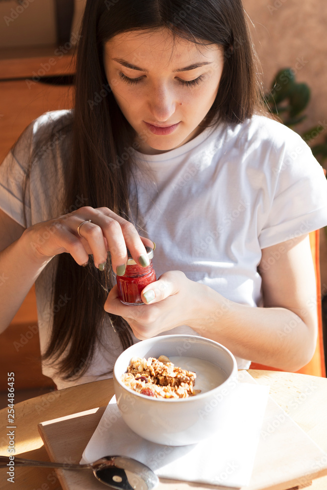 A young beautiful woman came to the cozy cafe in the morning to eat breakfast with a delicious granola sitting at a table near a large bright window