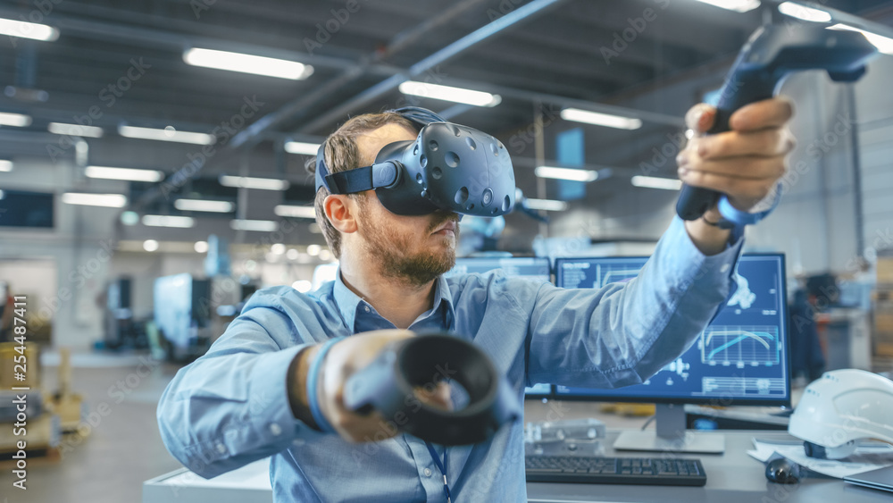 Portrait Shot of the Industrial Engineer Wearing Virtual Reality Headset and Using Controllers, ready to Work. In the Background Manufacturing Plant and Monitors.