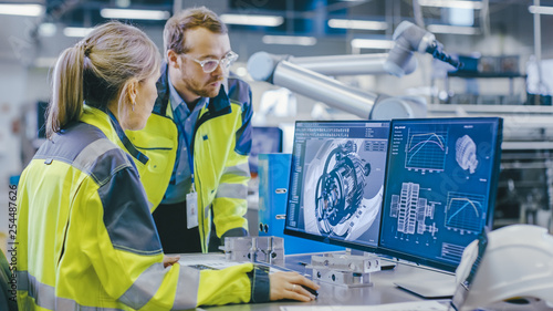 Schilderij op canvas At the Factory: Male Mechanical Engineer Holds Component while Female Chief Engineer Work on Personal Computer, They Discuss Details of the 3D Engine Model Design