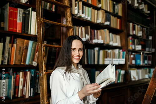 Young attractive student girl reading a book between library bookshelves