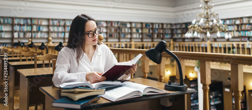 Frustrated female student sitting at the desk with a huge pile of study books in university library and studying