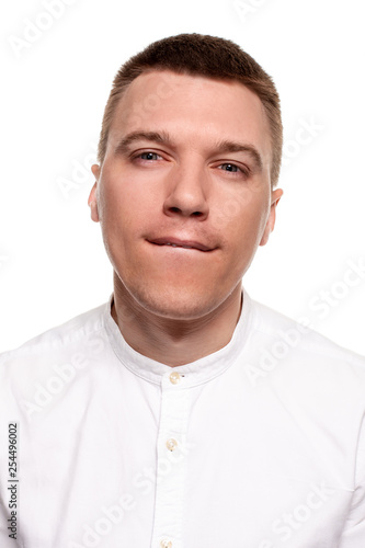 Charming handsome young man in a white shirt is making faces, while standing isolated on a white background