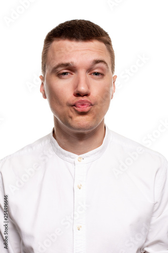 Charming handsome young man in a white shirt is making faces, while standing isolated on a white background