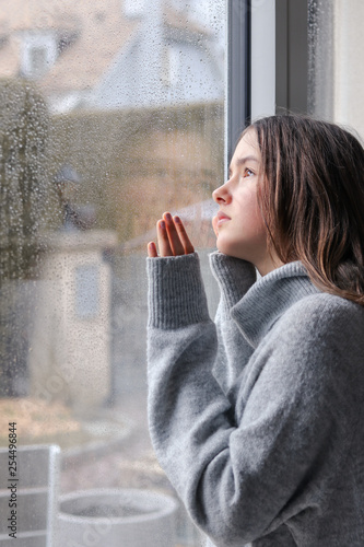 Картината върху платно Beautiful melancholic teenager girl in warm grey sweater looking outside through raindrops on wet window at sky at spring rainy day