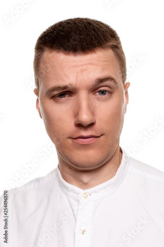 Charming handsome young man in a white shirt is making faces, while standing isolated on a white background