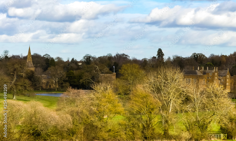 Landscape of Oxfordshire, with distant views of Broughton Castle, its gatehouse, moat  and associated church. Parkland with trees . Spring sunlight on buildings. Blue sky with white clouds. England.
