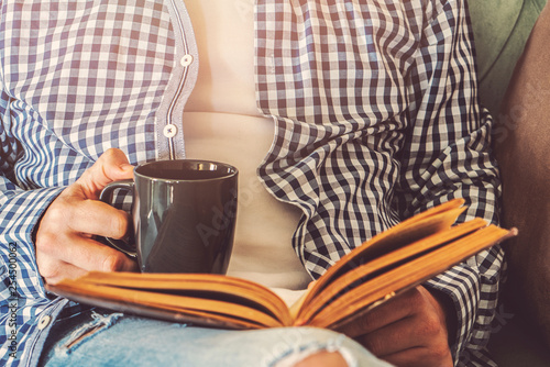 Young man is sitting on a sofa and reading a book while holding a cup of coffee or tea