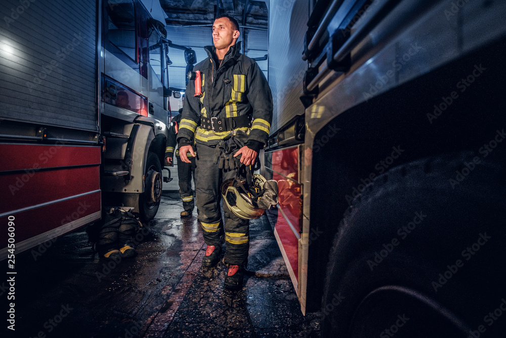 Full-length portrait of two brave firemen in protective uniform walking between two fire engines in the garage of the fire station