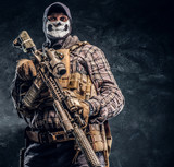 Private security service contractor wearing a balaclava skull and cap holding an assault. Studio photo against a dark textured wall