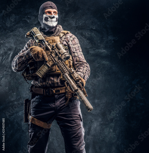 Private security service contractor wearing a balaclava skull holding an assault. Studio photo against a dark textured wall