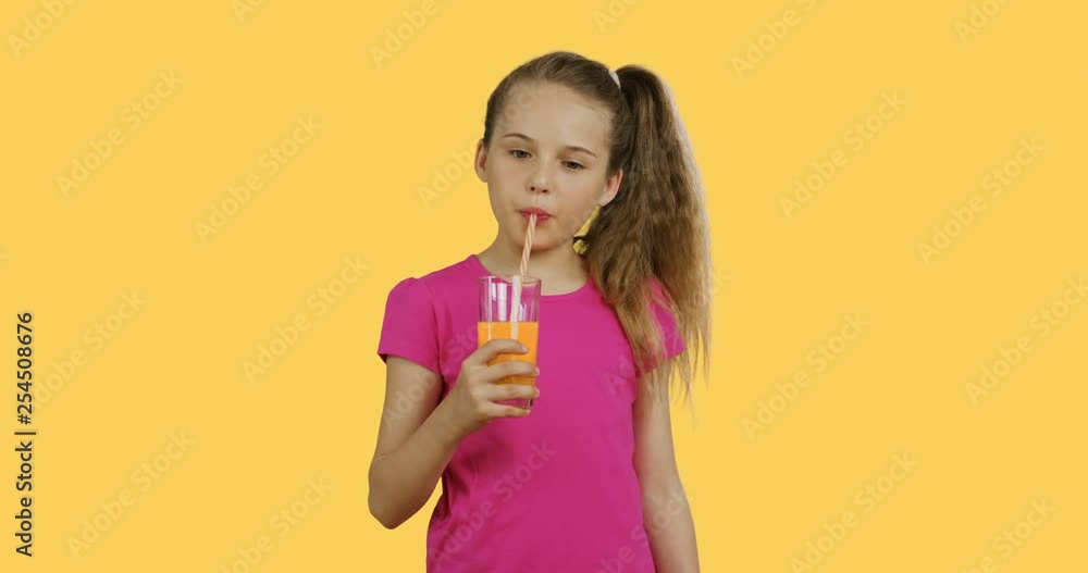 Cute girl with long hair in a tail in the pink T-shirt drinking orange juice from a glass through the straw on the yellow background. Cute girl with long hair in a tail in the pink T-shirt drinking orange juice from a glass through the straw on the yellow background.