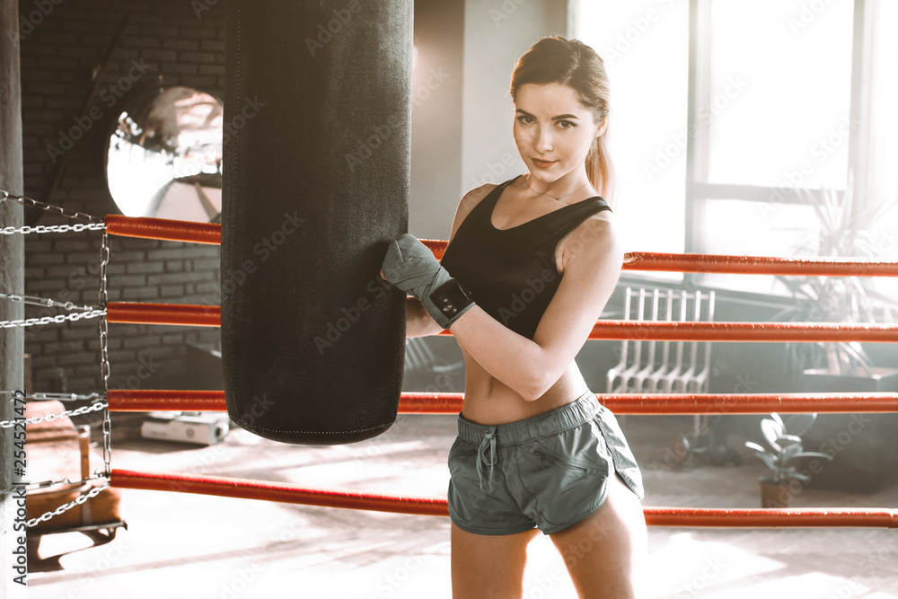 Female boxer training inside a boxing ring. Boxer practicing her moves at a boxing studio. The girl hugs a punching bag and looks at the camera
