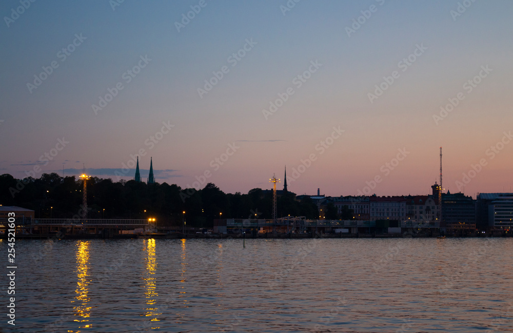 View of the silhouettes of the city of Helsinki with lights on the embankment and the spiers of cathedrals from the Gulf of Finland at dusk on a bright summer night in the capital of Finland. Backgrou