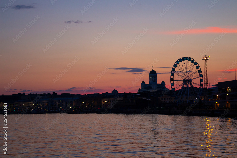 View of the embankment of Helsinki, the Ferris wheel, lights and the silhouette of the Cathedral of St. Nicholas from the Gulf of Finland at dusk on a bright summer night in the capital of Finland.