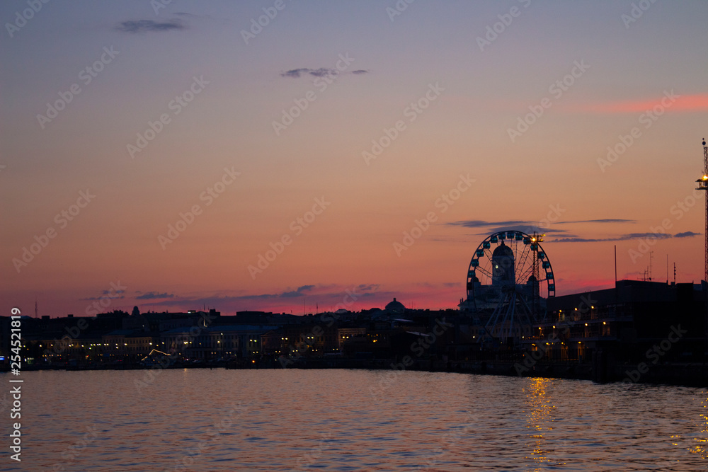 View of the embankment of Helsinki, the Ferris wheel, lights and the silhouette of the Cathedral of St. Nicholas from the Gulf of Finland at dusk on a bright summer night in the capital of Finland.