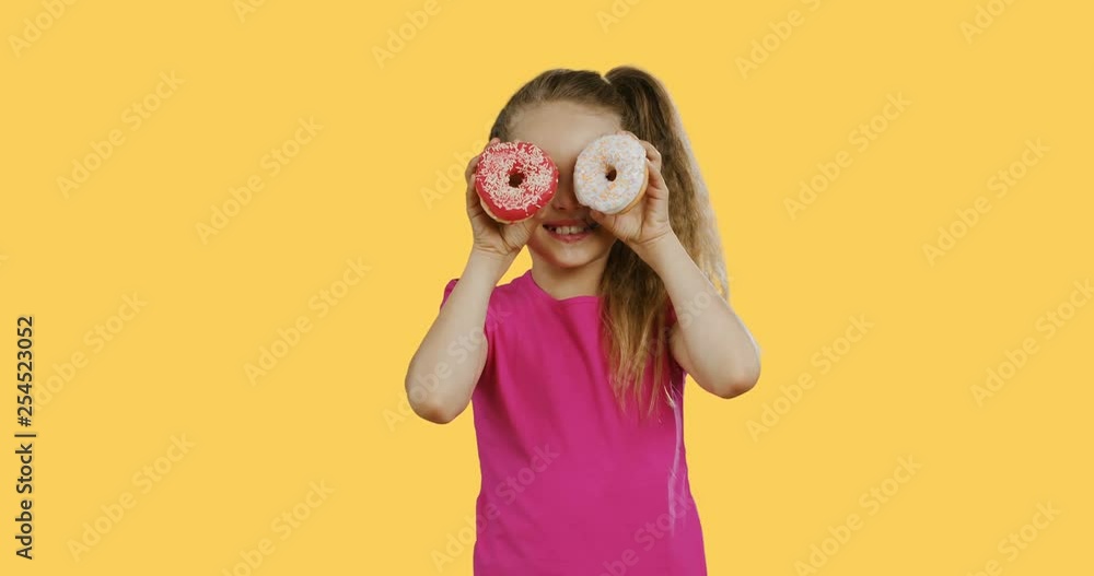 Pretty smiled teen girl in the pink T-shirt closing her face with donuts and smiling. Yellow wall background. Pretty smiled teen girl in the pink T-shirt closing her face with donuts and smiling. Yellow wall background.