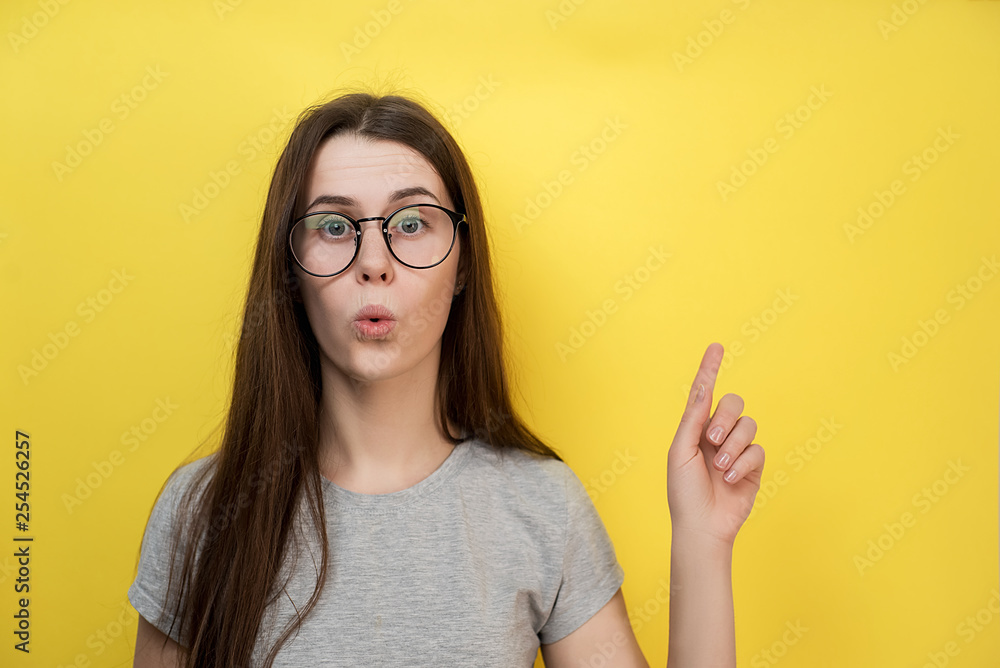 Surprised Caucasian young lady has bated breath, dressed in gray T-shirt, models against yellow background, demonstrates something at blank copy space for advertisement. Surprised Caucasian young lady has bated breath, dressed in gray T-shirt, models against yellow background, demonstrates something at blank copy space for advertisement.