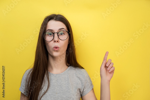 Surprised Caucasian young lady has bated breath, dressed in gray T-shirt, models against yellow background, demonstrates something at blank copy space for advertisement.