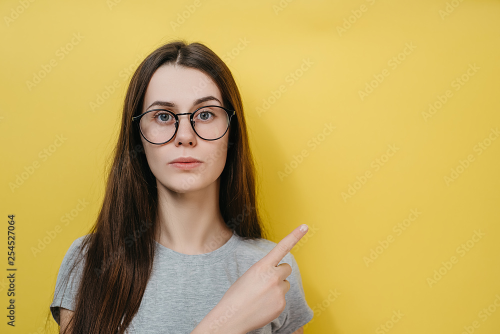 Young female in glasses with has bated breath, stares at camera, attracts your attention at copy space aside, demonstrates some items in shop, isolated over yellow wall Young female in glasses with has bated breath, stares at camera, attracts your attention at copy space aside, demonstrates some items in shop, isolated over yellow wall