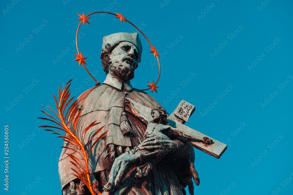 Naklejka premium Baroque Statue of Saint John Nepomuk with golden star halo holding crucifix with Jesus on Charles Bridge. Prague, Czech Republic. Orange and teal mood
