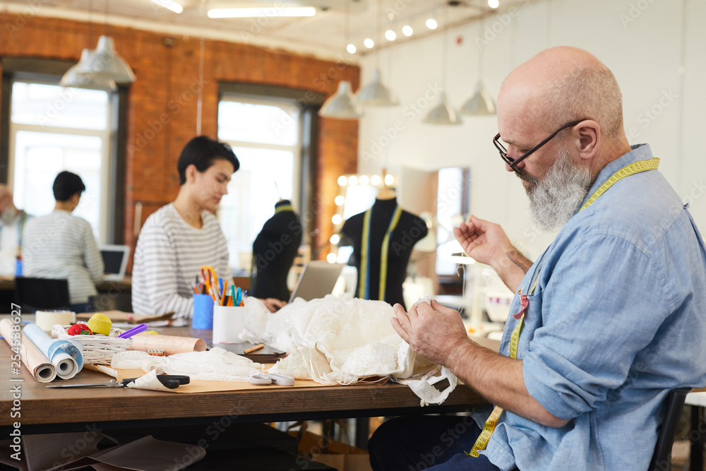 Mature grey-bearded man in shirt sitting by table and sewing white dress or other item for client in workshop Mature grey-bearded man in shirt sitting by table and sewing white dress or other item for client in workshop