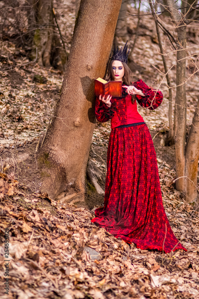 Art Photography. Gorgeous Mysterious Fairy Princess in Red Dress and Black Crown With Old Book. Posing in Forest Outdoors.