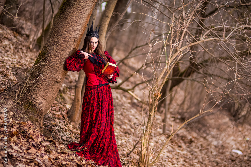 Art Photography. Gorgeous Mysterious Fairy Princess in Red Dress and Black Crown With Old Book. Posing in Forest Outdoors.