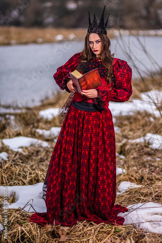 Art Photography Ideas. Mysterious Fairy Princess in Red Dress and Black Crown Reading Old Book. Posing in Forest Outdoors.