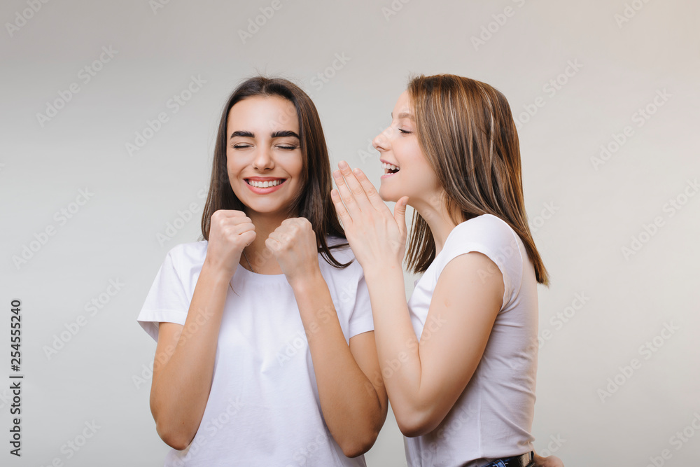 Two lovely women gossiping while one is telling something to another while another is laughing with closed eyes being happy against a grey wall.. Two women gossiping secrets Two lovely women gossiping while one is telling something to another while another is laughing with closed eyes being happy against a grey wall.. Two women gossiping secrets