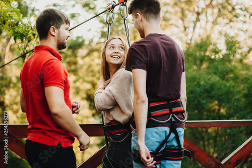 Young lovely couple being equipped before rope slide while girl is looking at her boyfriend laughing with fear while rope slide worker is looking at them smiling.