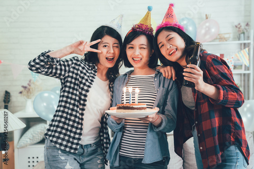 Group of young friends having a birthday party in decorated room home. charming girls face camera holding cake with lighted candles and drinking beer blinking eyes smiling. beautiful ladies celebrate