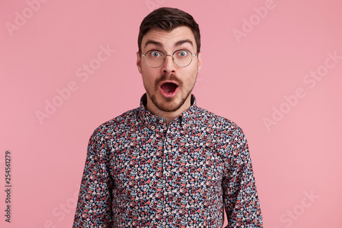 Shocked attractive dark-haired handsome young man staring throught glasses, unshaved with beard and mustache in colorful shirt opened mouth from surprise, isolated over pink background photo